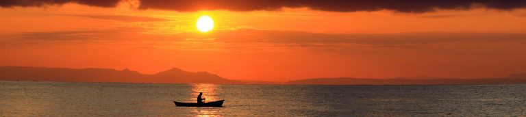 A view of the ocean with a small boat and the sun setting behind it.